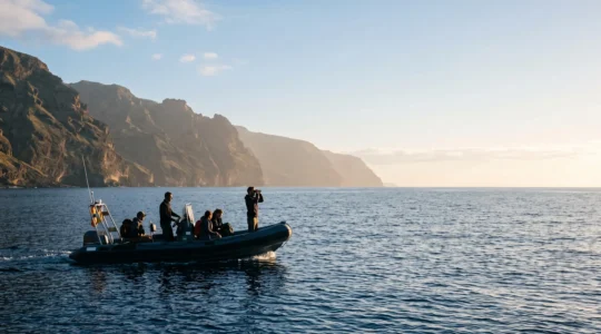 Petit bateau d'observation scientifique naviguant au large des falaises volcaniques des Canaries au lever du soleil, silhouettes de participants scrutant l'horizon